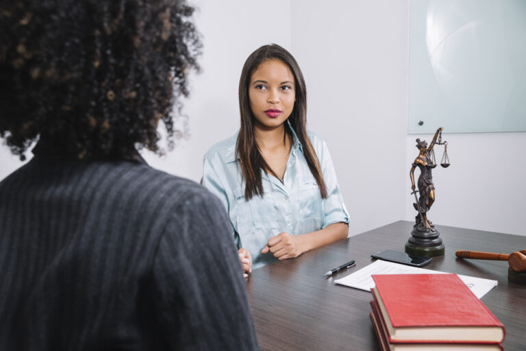 Two women talking across a desk beside a statue of the scales of justice and a gavel