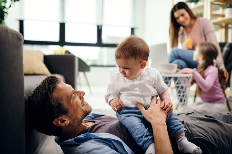 A father playing with his baby son on the floor while the mother and daughter talk nearby